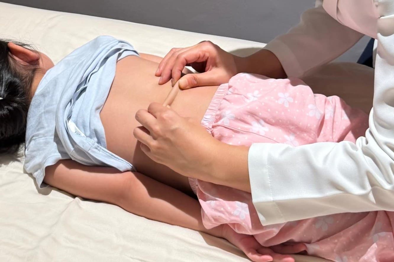 A physician working along a child's back with a small wooden tui na tool, the child lying face down.